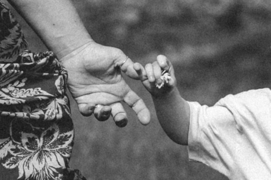 Black and white close-up of an adult holding a small child's hand, with the child grasping the adult's finger outdoors.
