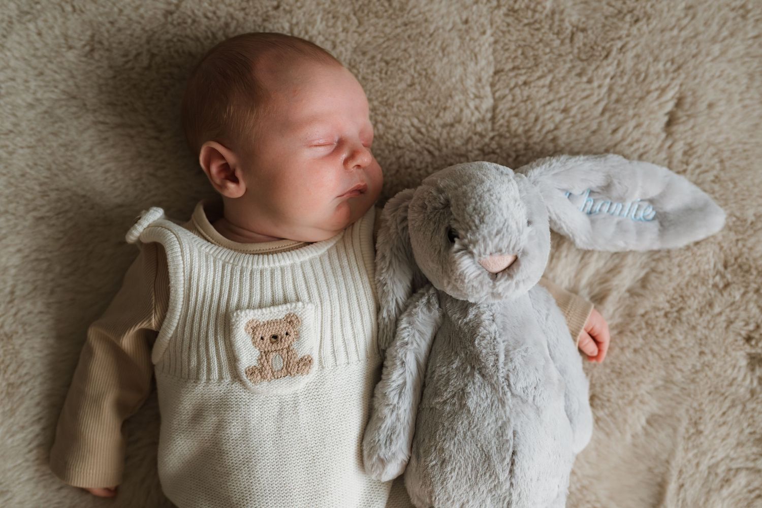 Newborn baby in a cream knit romper with a teddy bear patch sleeping beside a soft grey bunny toy on a fluffy blanket.