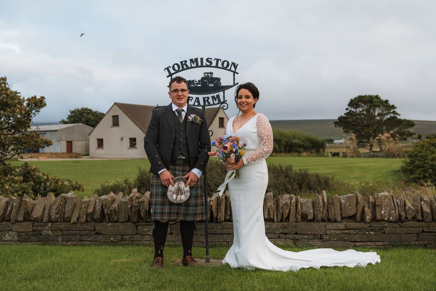 Smiling bride and groom in a tartan kilt pose with a bouquet on a lawn in front of a stone wall and a farm sign.