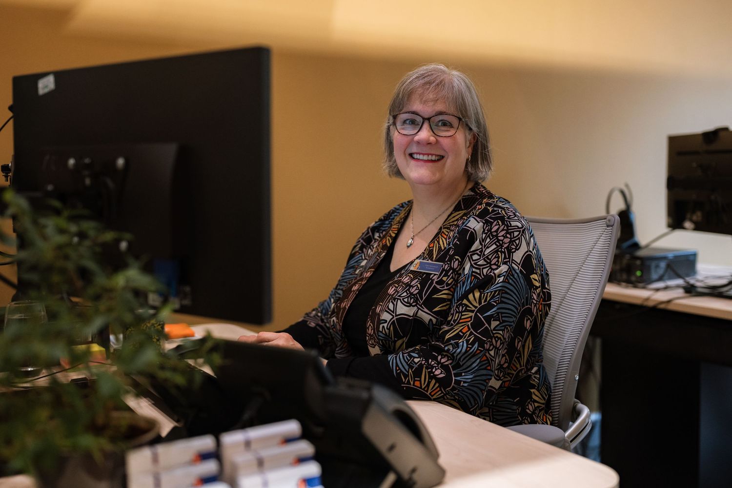Smiling woman with glasses and a patterned blouse sits at a desk with computer monitors in a bright, modern office.