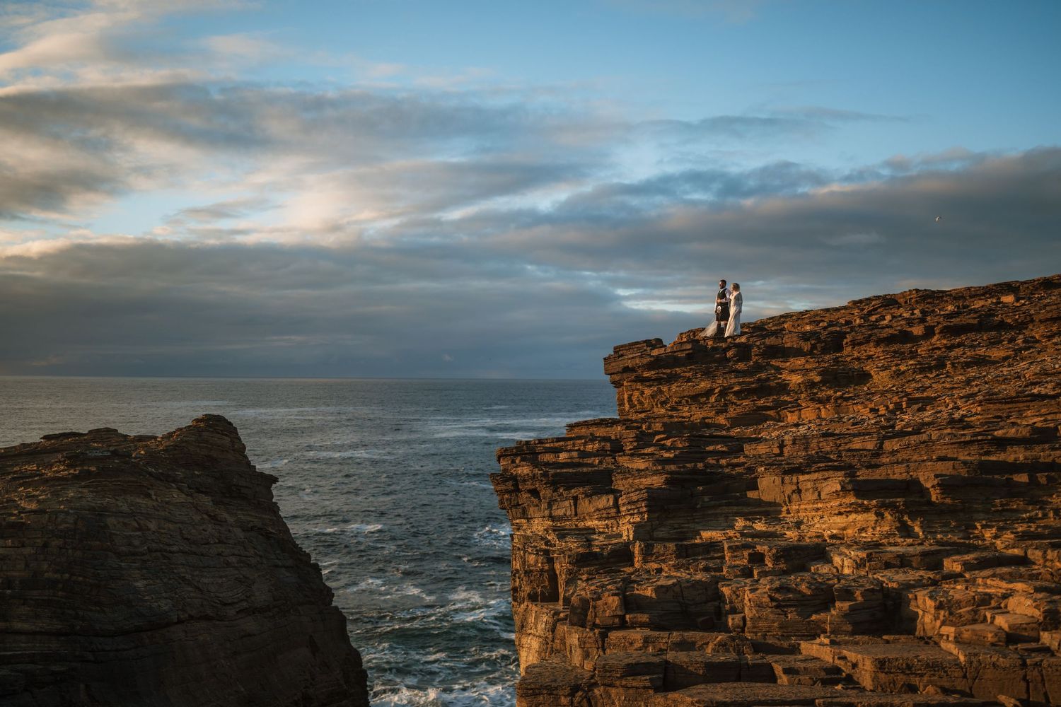 Tiny wedding couple stands on a dramatic sea cliff edge beneath a colorful sky with layered rock formations and ocean below.