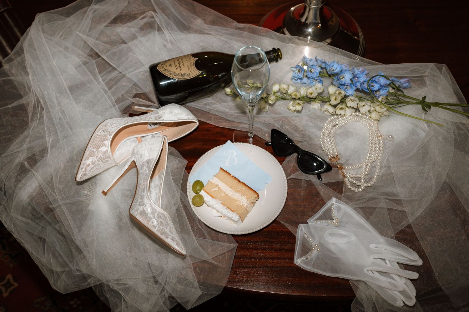 Wedding details including heeled shoes, lace gloves, pearls, sunglasses, champagne and a cake slice arranged on a table with tulle.