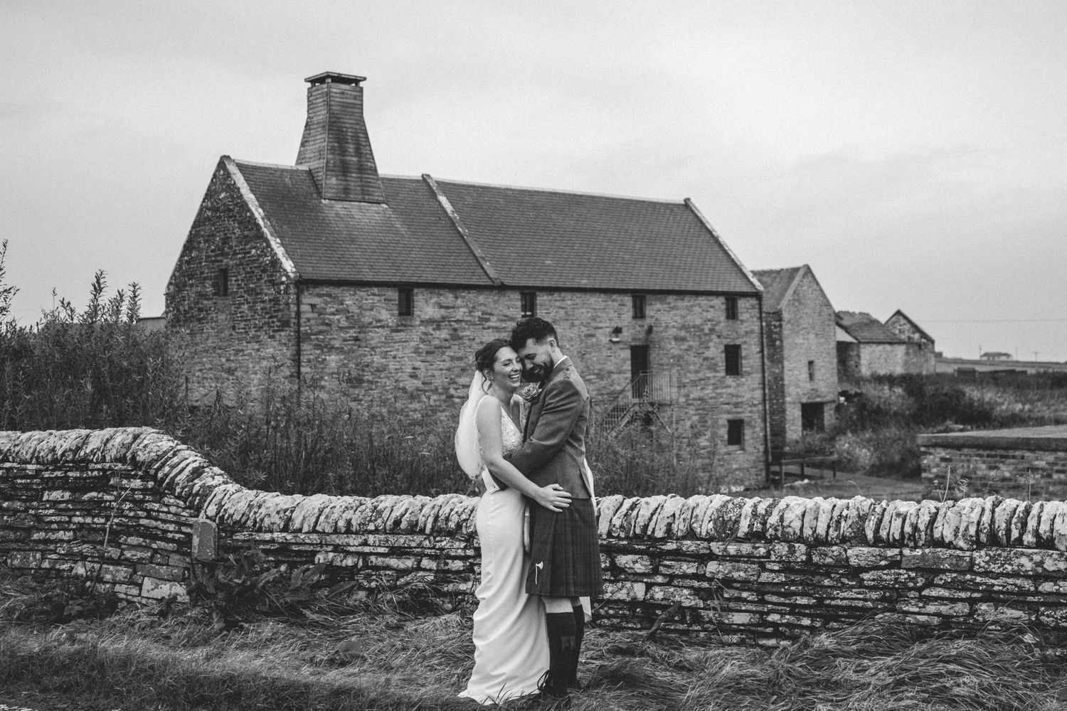 Bride and groom in a kilt embrace beside a dry stone wall in front of a historic stone building in black and white.