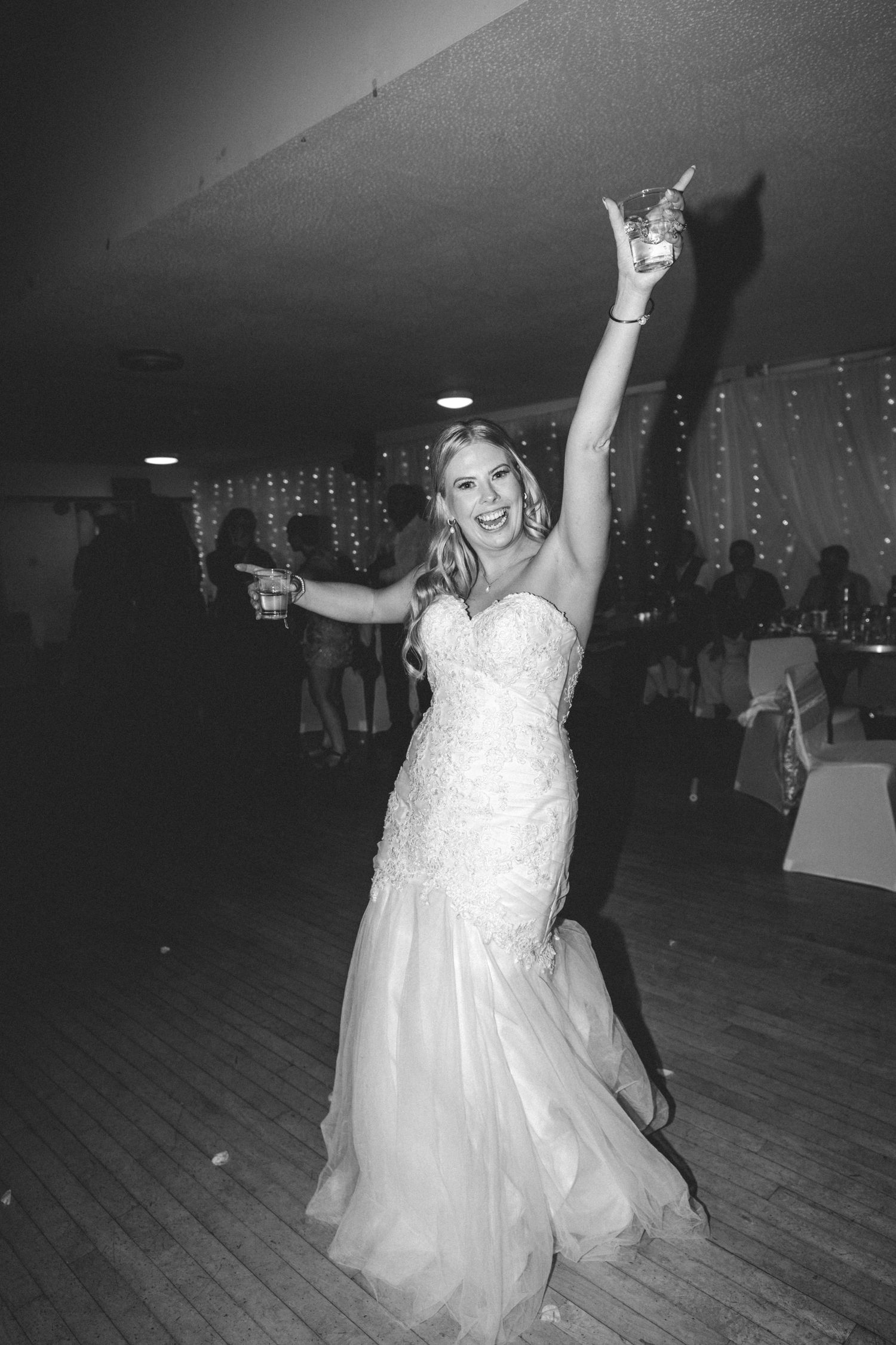 Joyful bride in a lace gown dances with her arms raised on a dance floor in black and white.