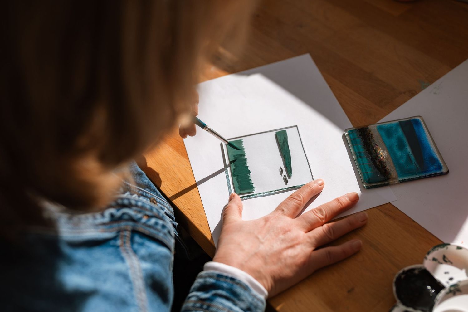 Woman in a denim jacket paints green and blue watercolor designs on small glass panels at a wooden table with paper and paint supplies.