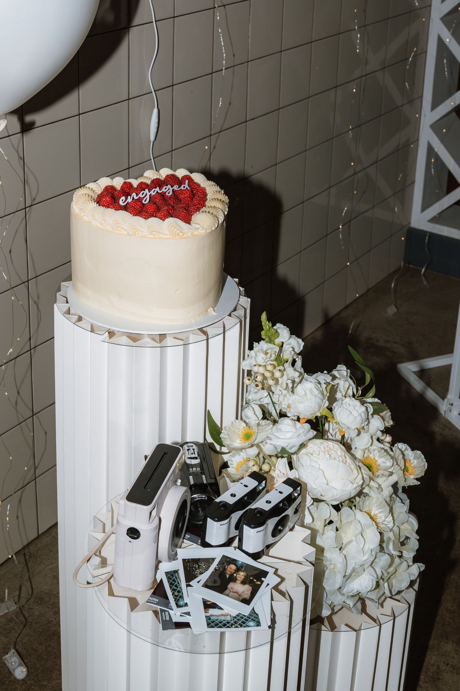 White engagement cake with red berries on a pleated pedestal beside white peonies, instant cameras and Polaroid photos.