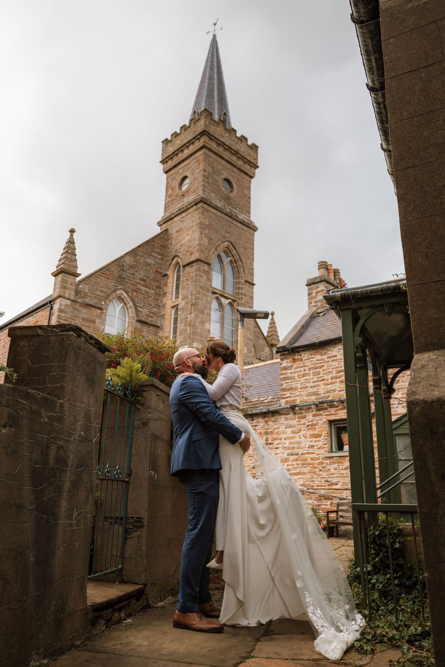 Bride and groom embrace in a stone courtyard with a historic church tower rising behind them.
