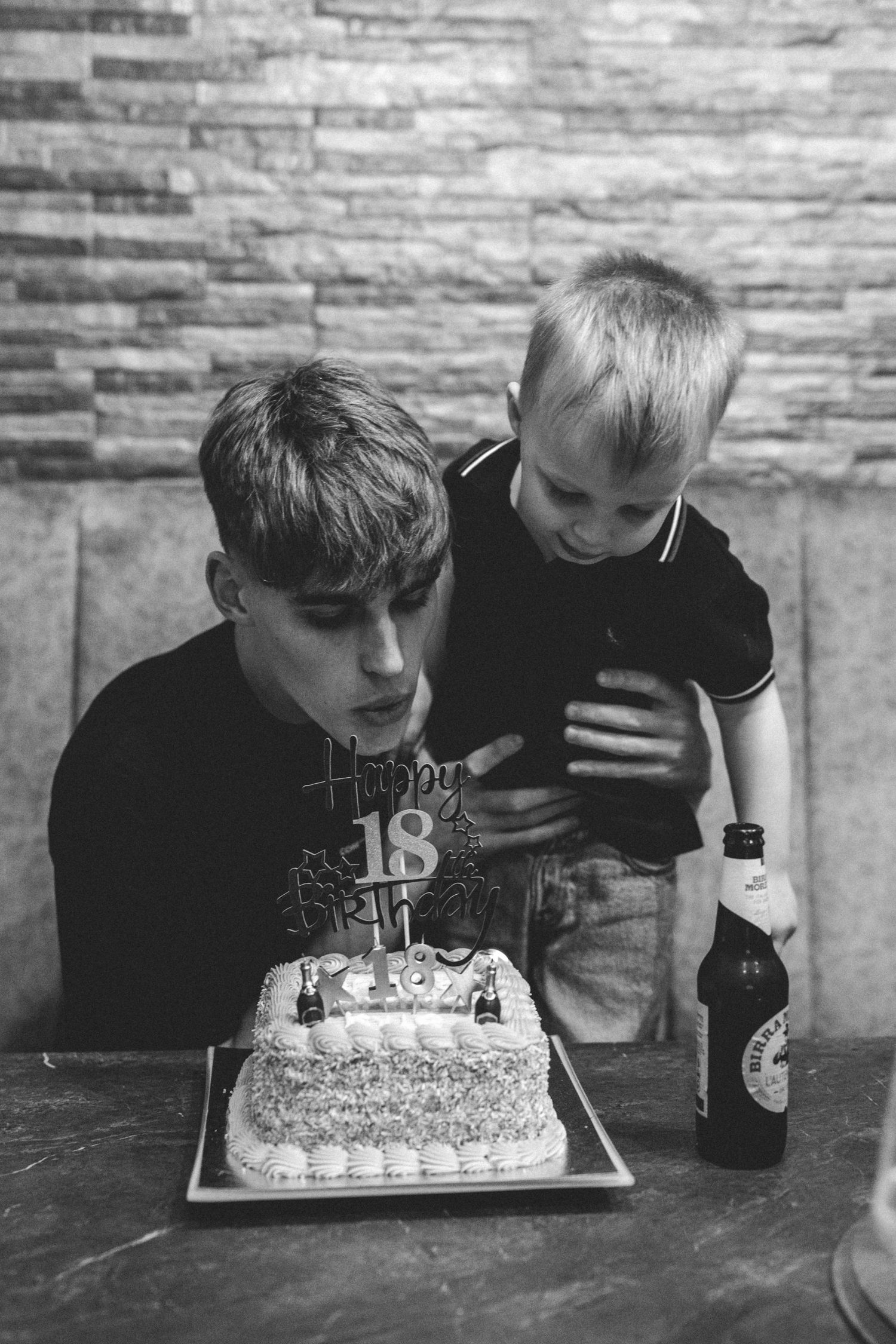 Young man blowing out candles on a Happy 18 birthday cake while a small boy leans on his shoulder, in black and white.