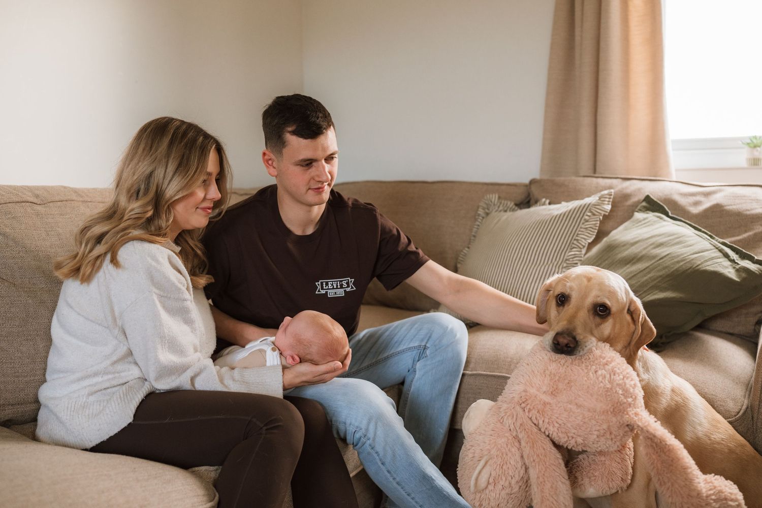 A smiling couple sits on a beige couch holding their newborn baby while a yellow Labrador rests its head beside them.