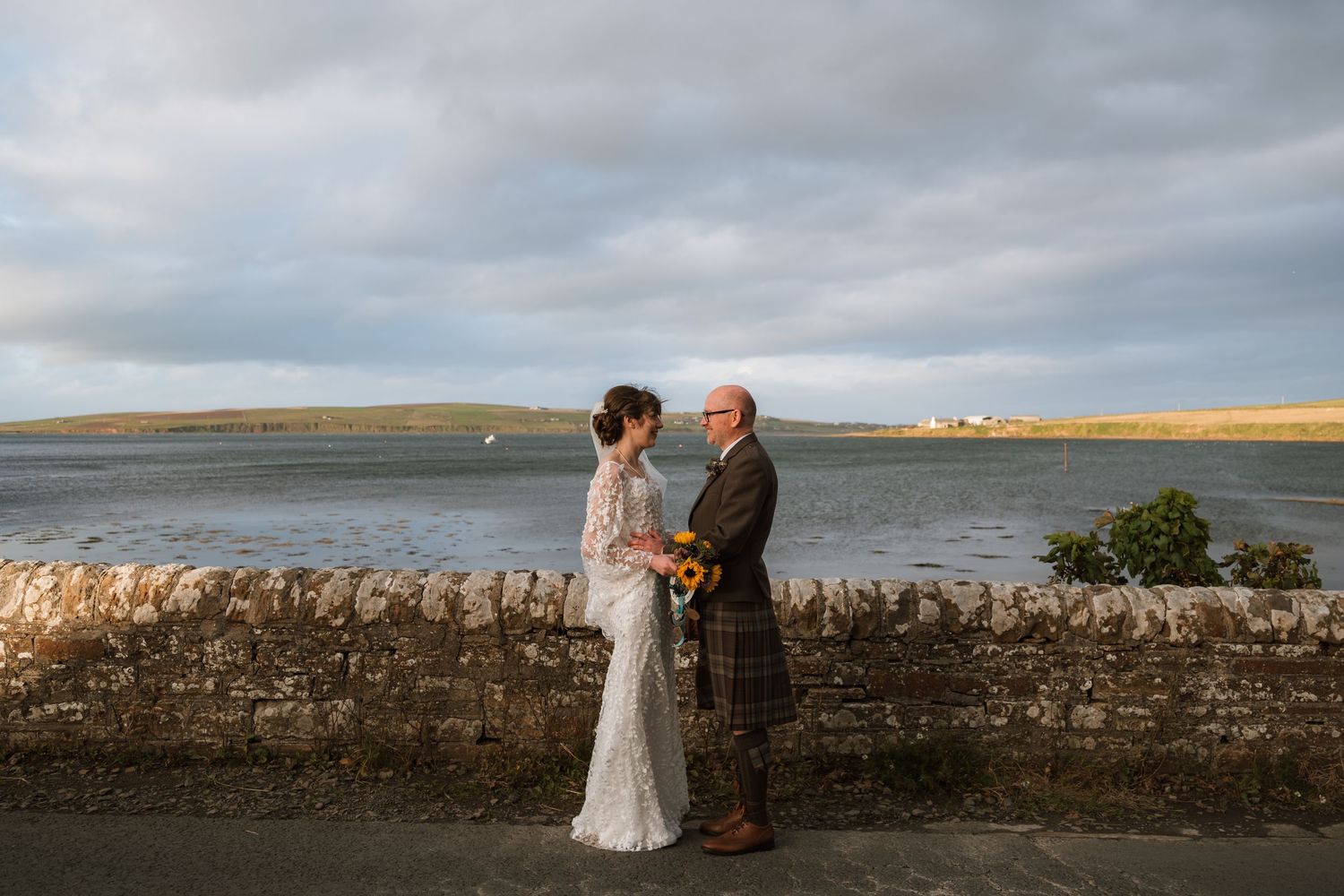 Bride in a lace gown and groom in a kilt hold hands beside a stone wall with a calm bay and green hills behind them.