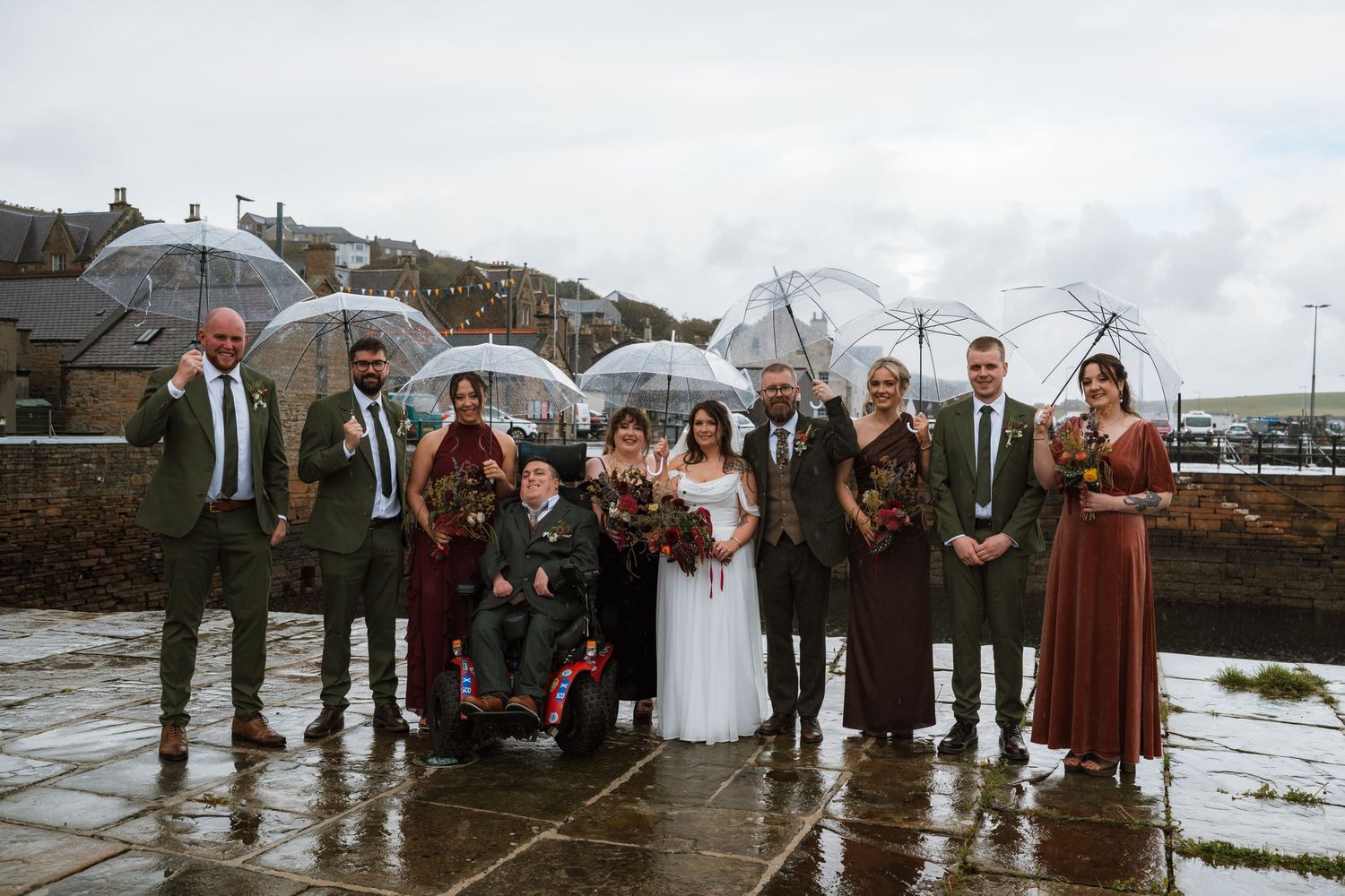Wedding party in formal attire gathers with a bride in white under clear umbrellas on a wet stone pavement by a coastal town.