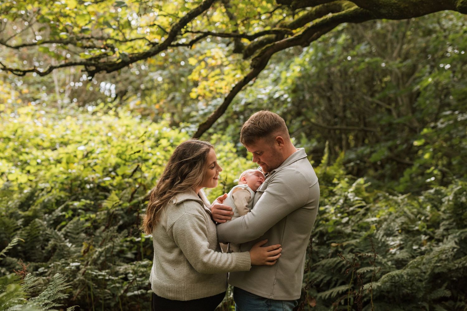 Couple embracing their newborn baby face to face among sunlit trees and ferns in a woodland family portrait.