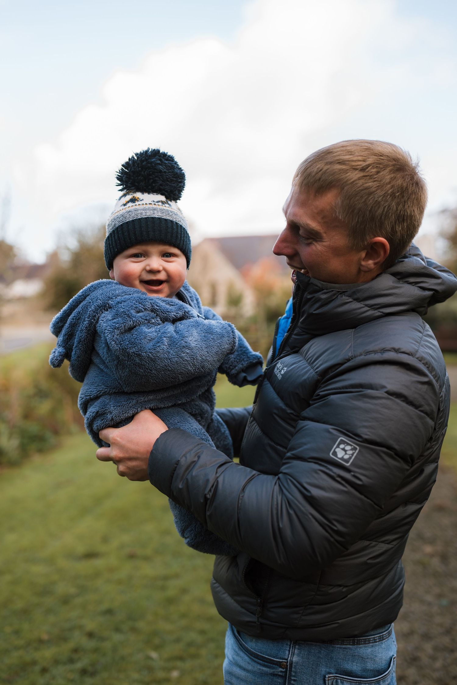 Smiling baby in a blue knit outfit and pom-pom beanie being held up by a man in a black puffer jacket on a grassy path outdoors.