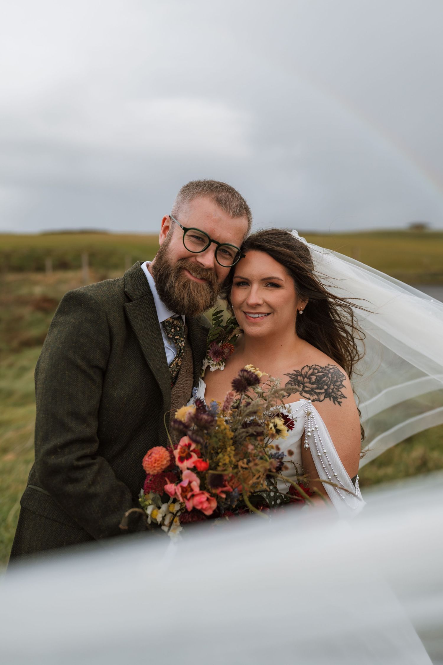Bearded groom in a tweed jacket and tattooed bride with a colorful bouquet embrace in a windswept field with veil blowing.