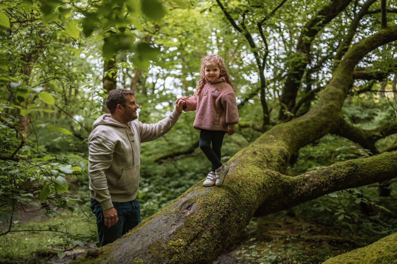 Father helps a little girl in a pink jacket balance along a mossy tree branch in the forest.