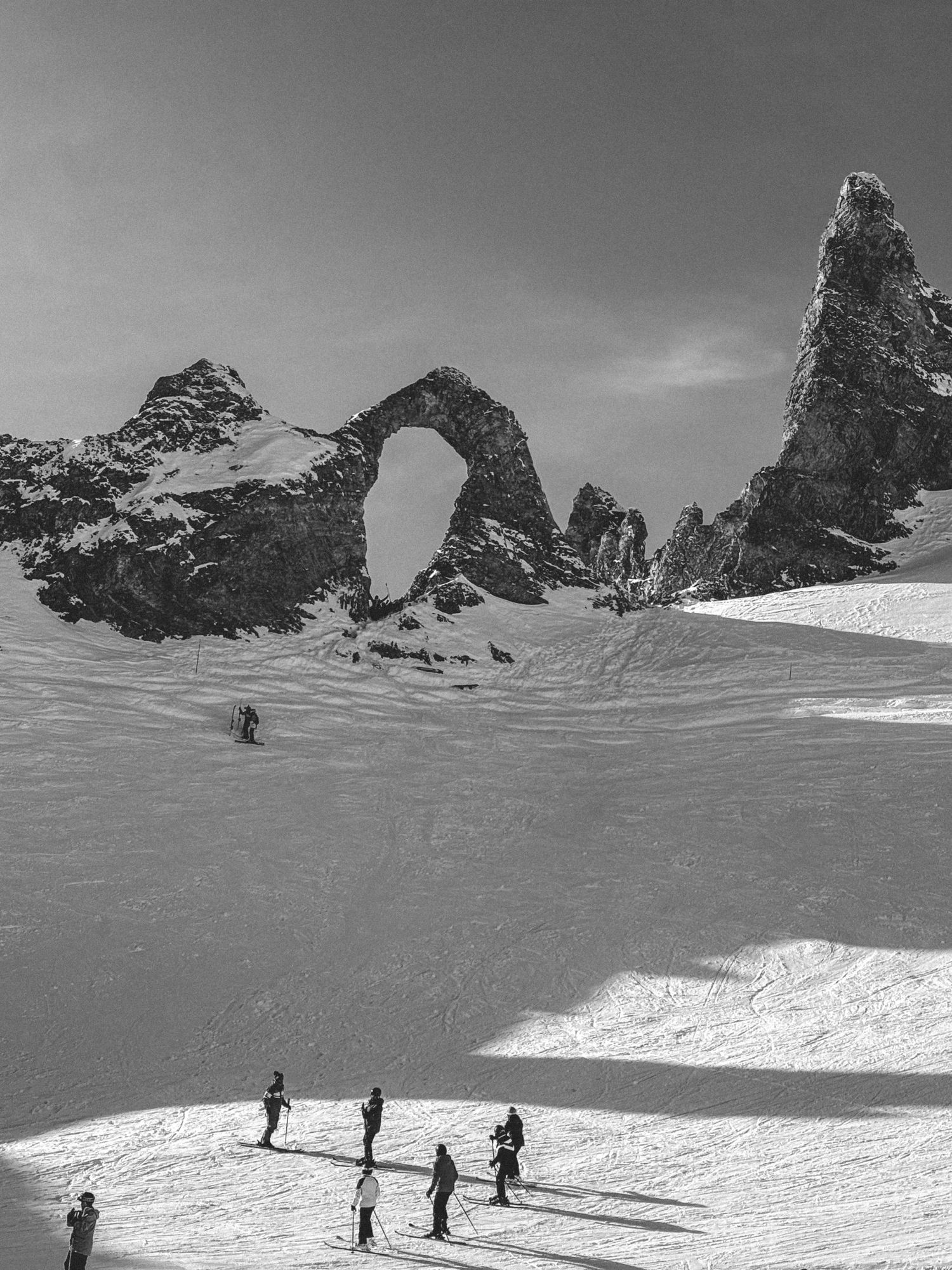 Black and white photo of skiers on a snowy slope below a rock arch and jagged mountain peaks.