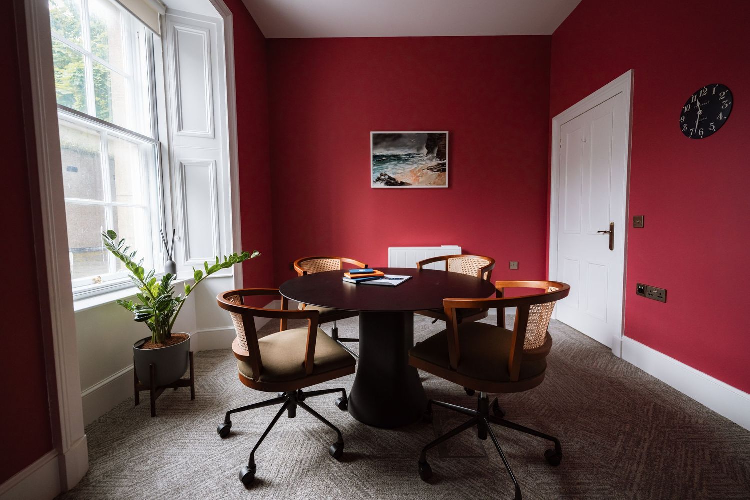 Cozy meeting room with deep red walls, a round dark wood table, wooden swivel chairs, a potted plant, and a wall clock.