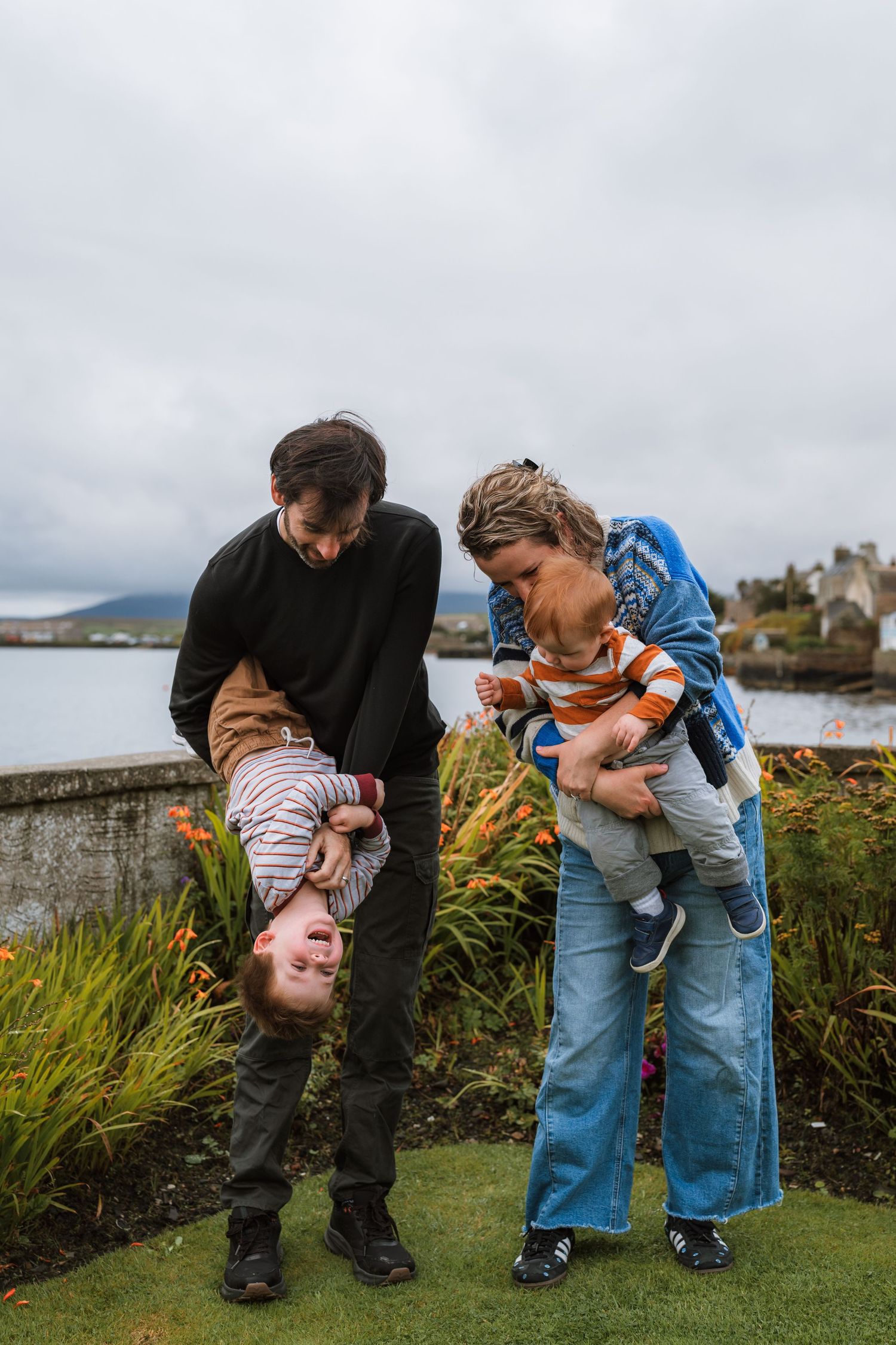 A father swings a laughing child upside down while a mother holds a toddler on a grassy lawn by the water.