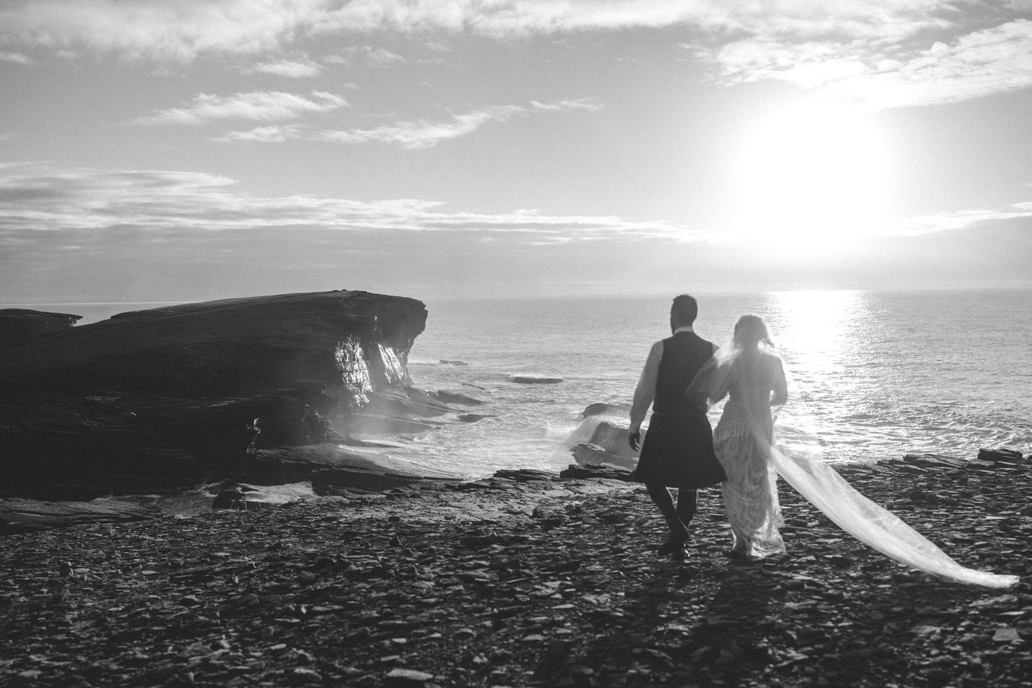 Black and white photo of a bride and groom walking along a rocky beach at sunset with the bride's veil trailing behind.