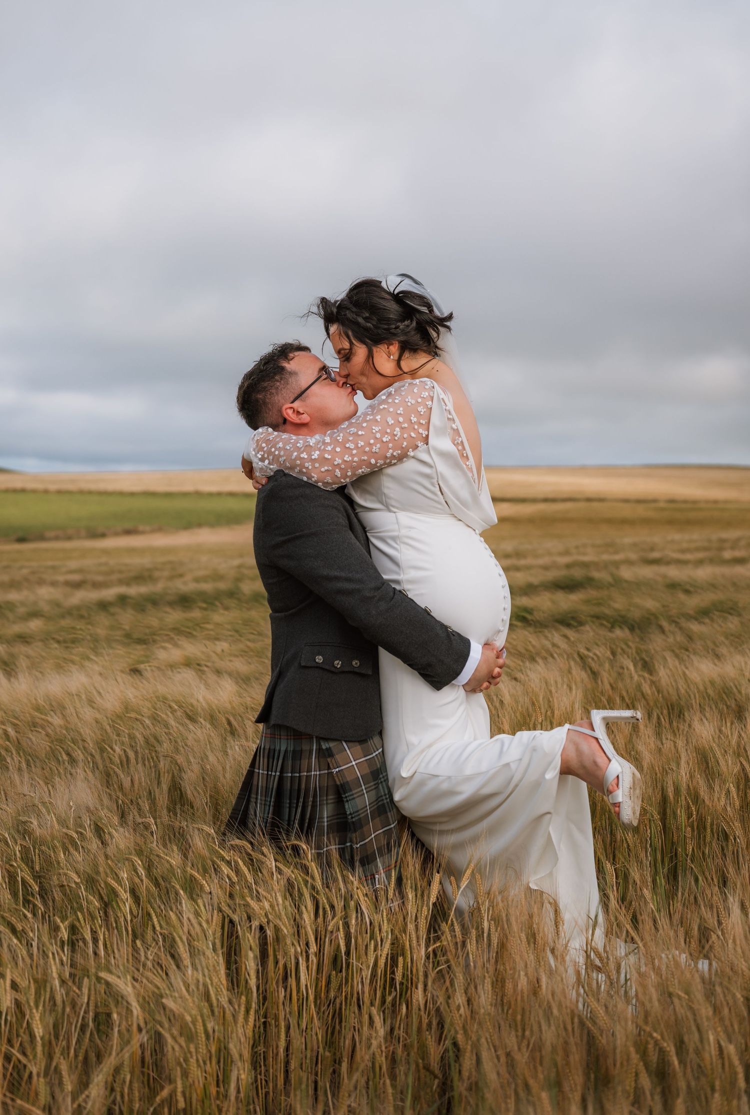 Groom in a kilt lifts and kisses his bride in a golden wheat field beneath a cloudy sky.
