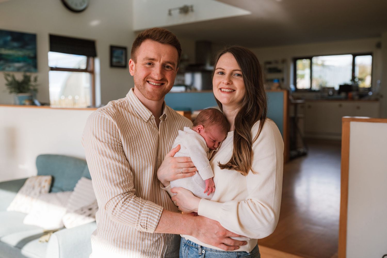 Smiling couple standing in a bright modern living room, gently cradling their sleeping newborn baby between them.
