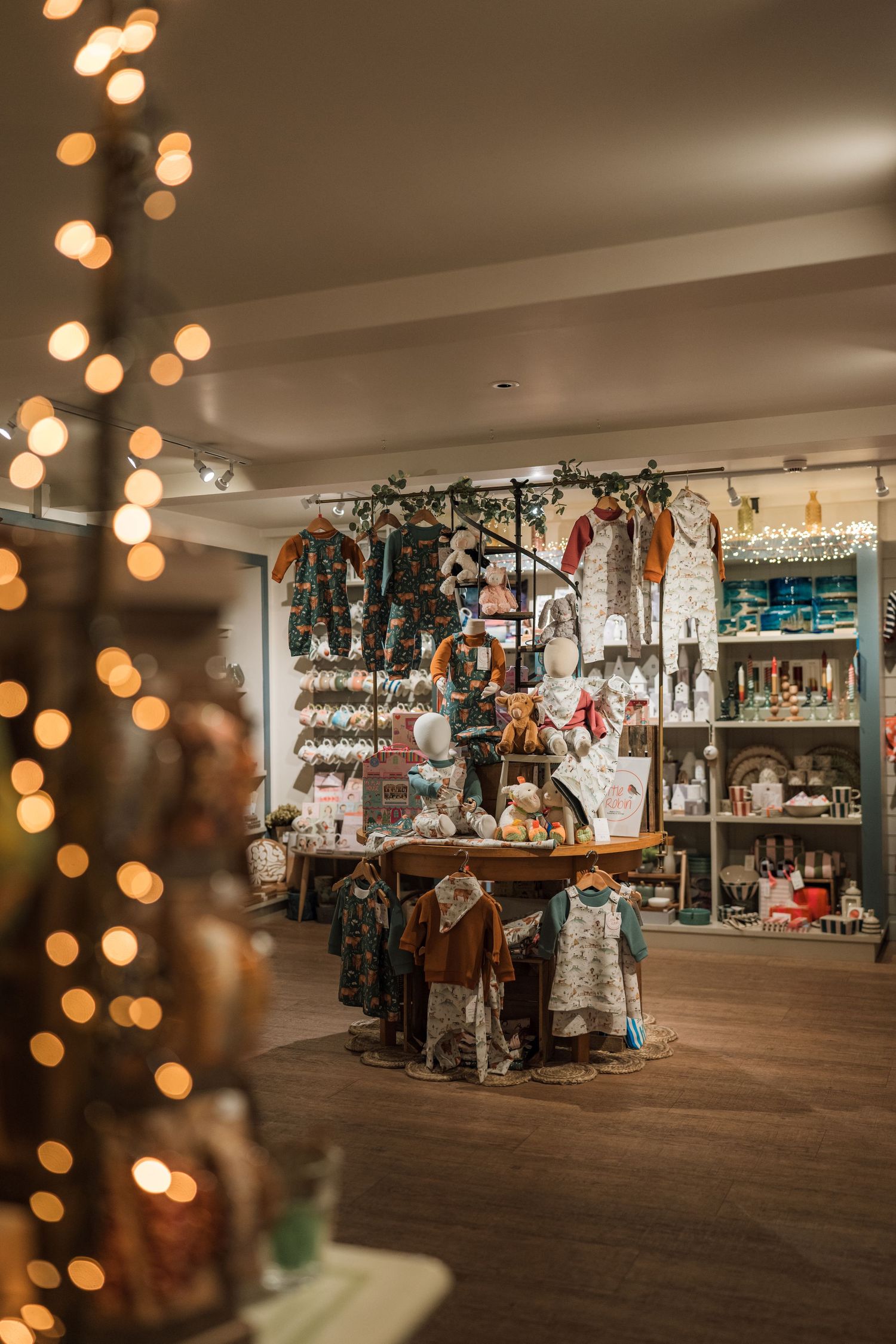 Cozy boutique interior with warm string lights, children's clothing on display tables and racks, and shelves of gifts and home decor.