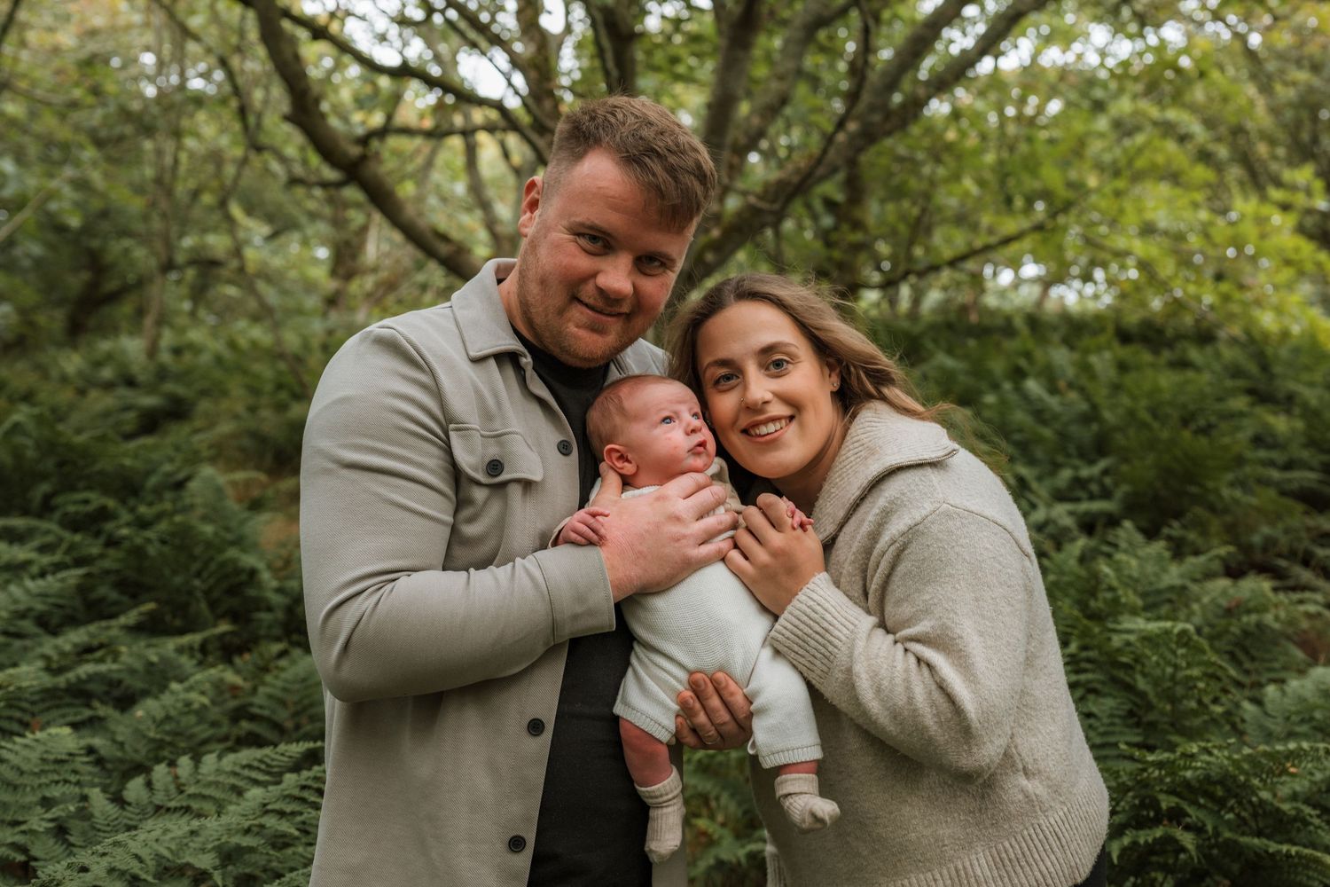Smiling couple cuddling their newborn baby between them in a lush green woodland during a family photoshoot.