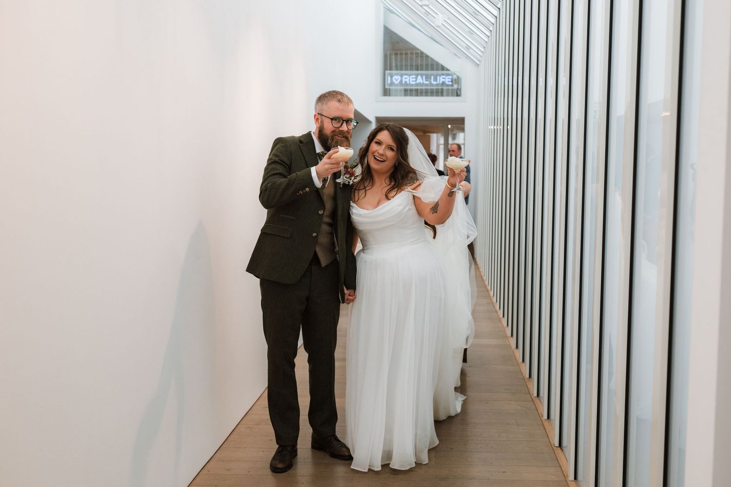 Smiling bride and groom raise glasses in a bright modern gallery corridor, celebrating their wedding day.
