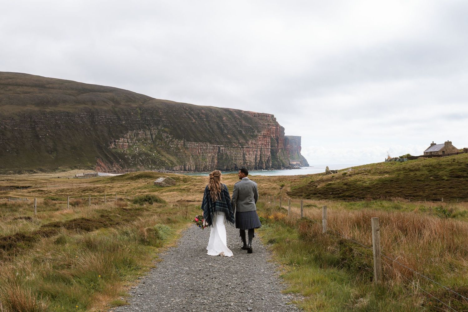 Bride and groom in a kilt walk down a gravel path toward towering sea cliffs on a moody Scottish coast.