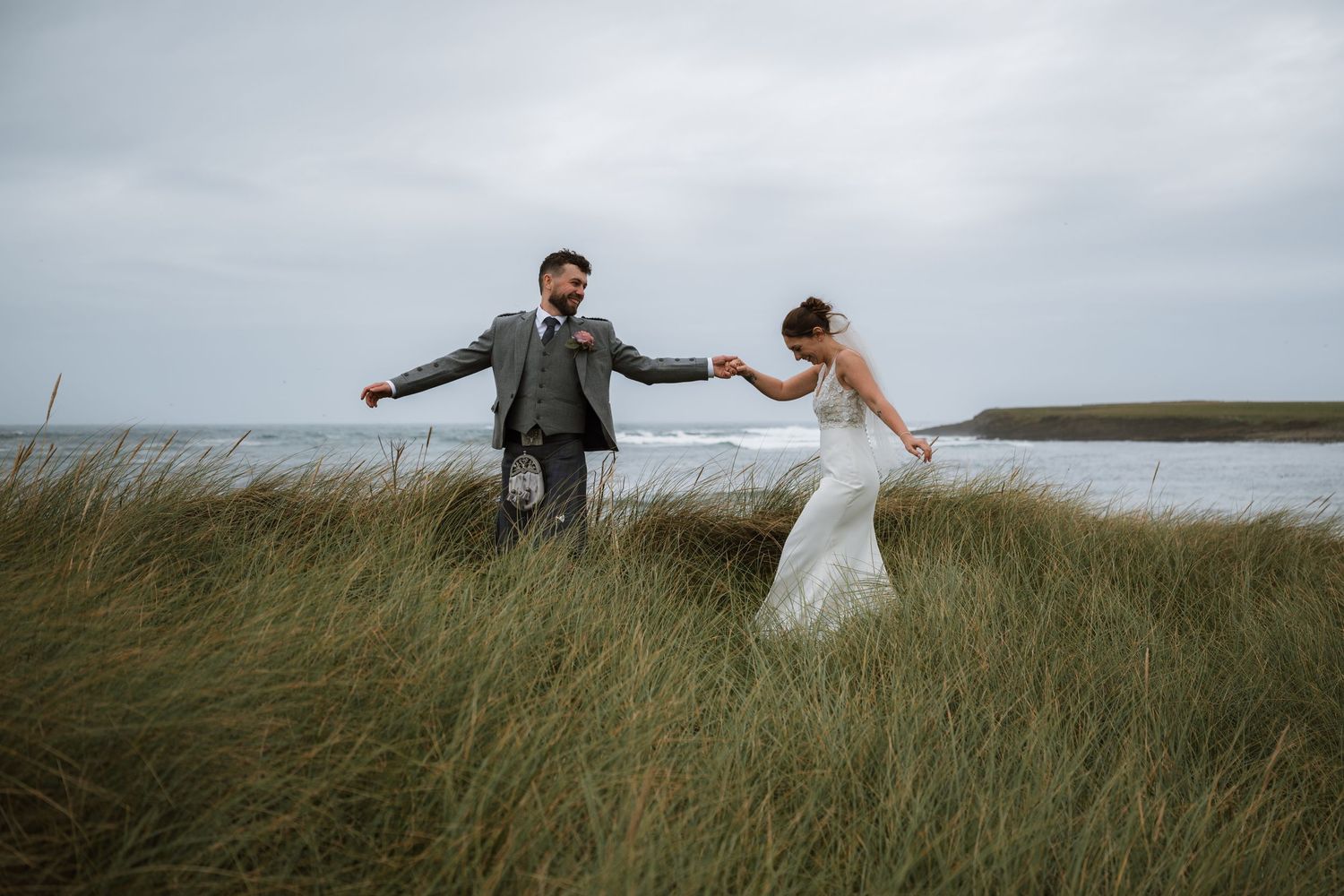 A bride and groom hold hands and dance in tall coastal grass beside the sea under a moody, overcast sky.