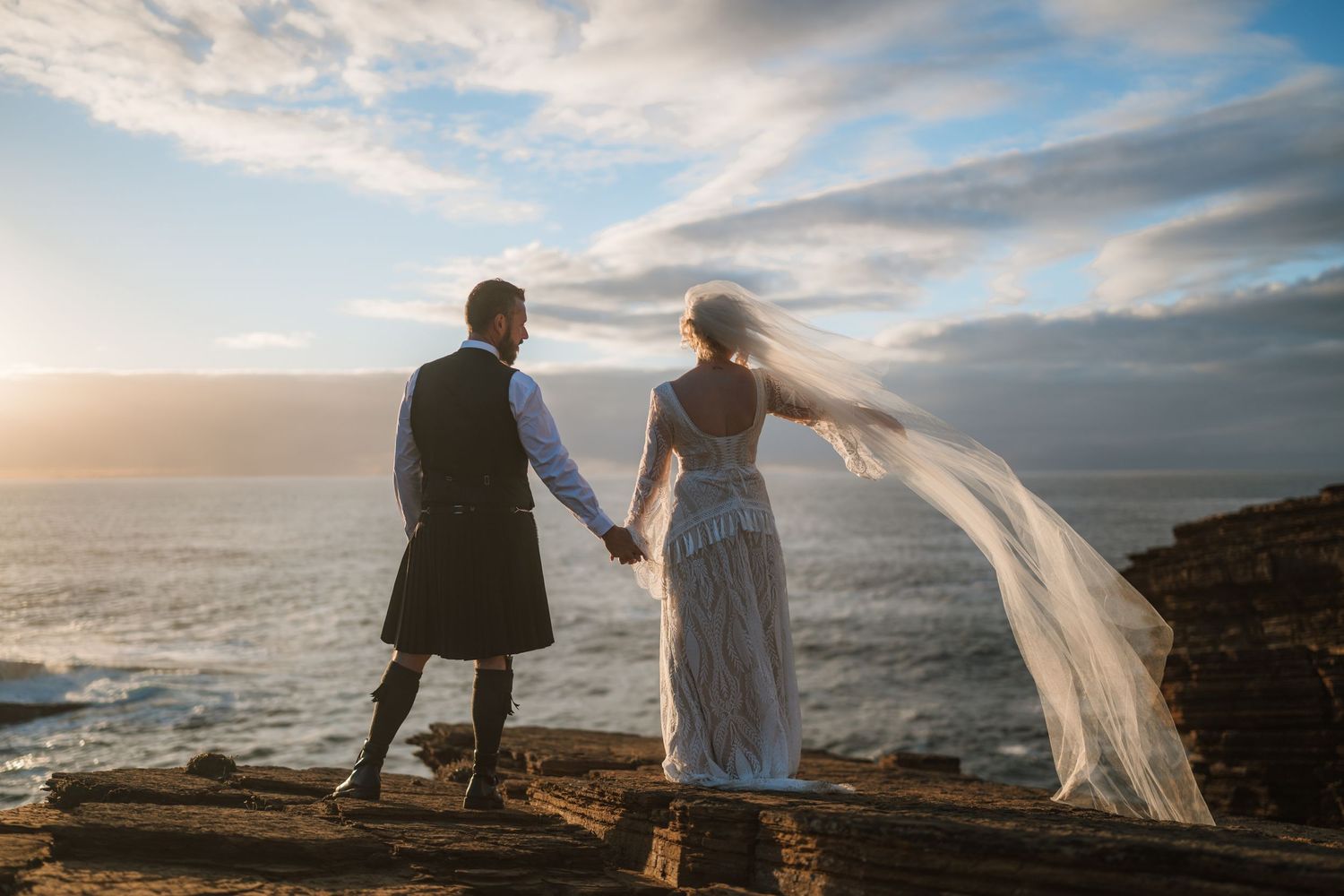 Bride in a flowing white gown and veil holds hands with a groom in a kilt on a rocky clifftop overlooking the sea at sunset.