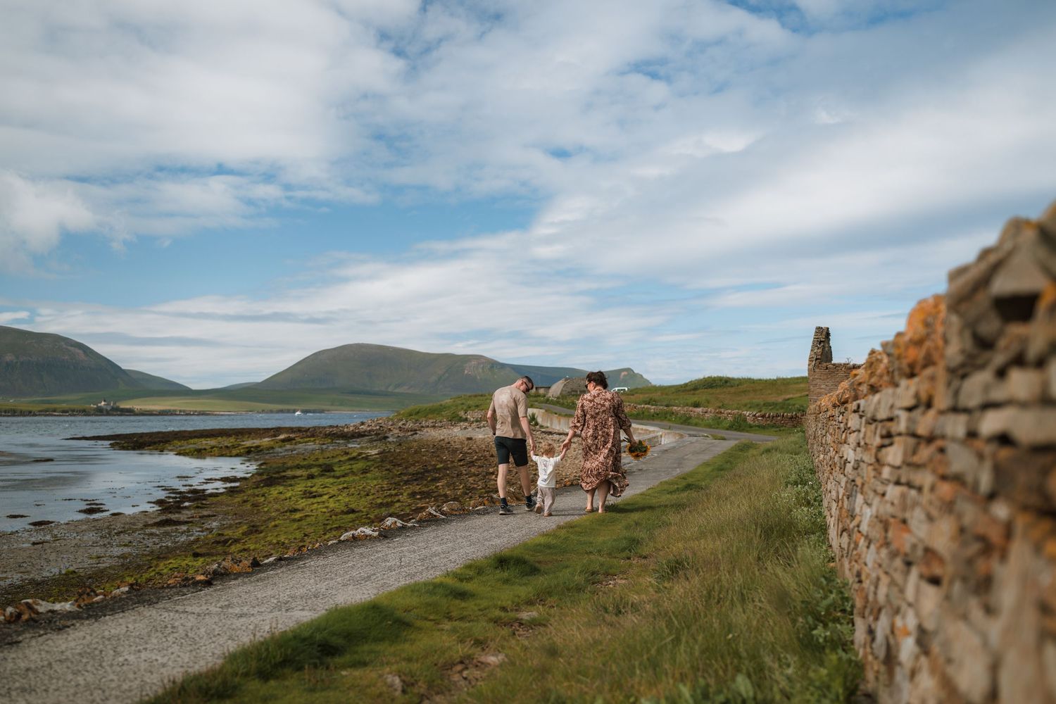A couple walks a dog along a coastal path beside a stone wall with calm water and rolling green hills in the background.