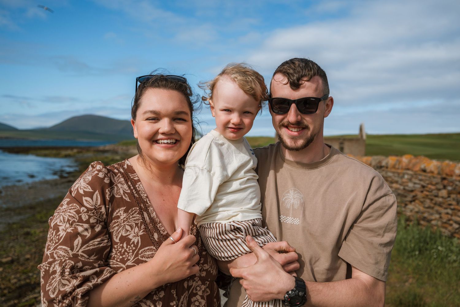 A smiling family of three, a woman, a man in sunglasses and a toddler, pose together outdoors by the sea and a stone ruin.