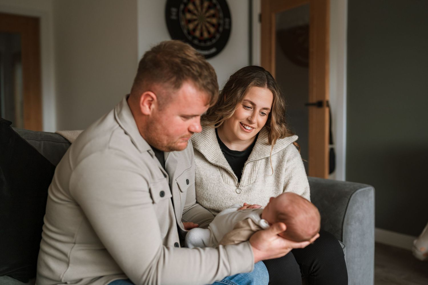 Smiling mother and father sit on a grey sofa at home, gazing lovingly at their sleeping newborn baby.
