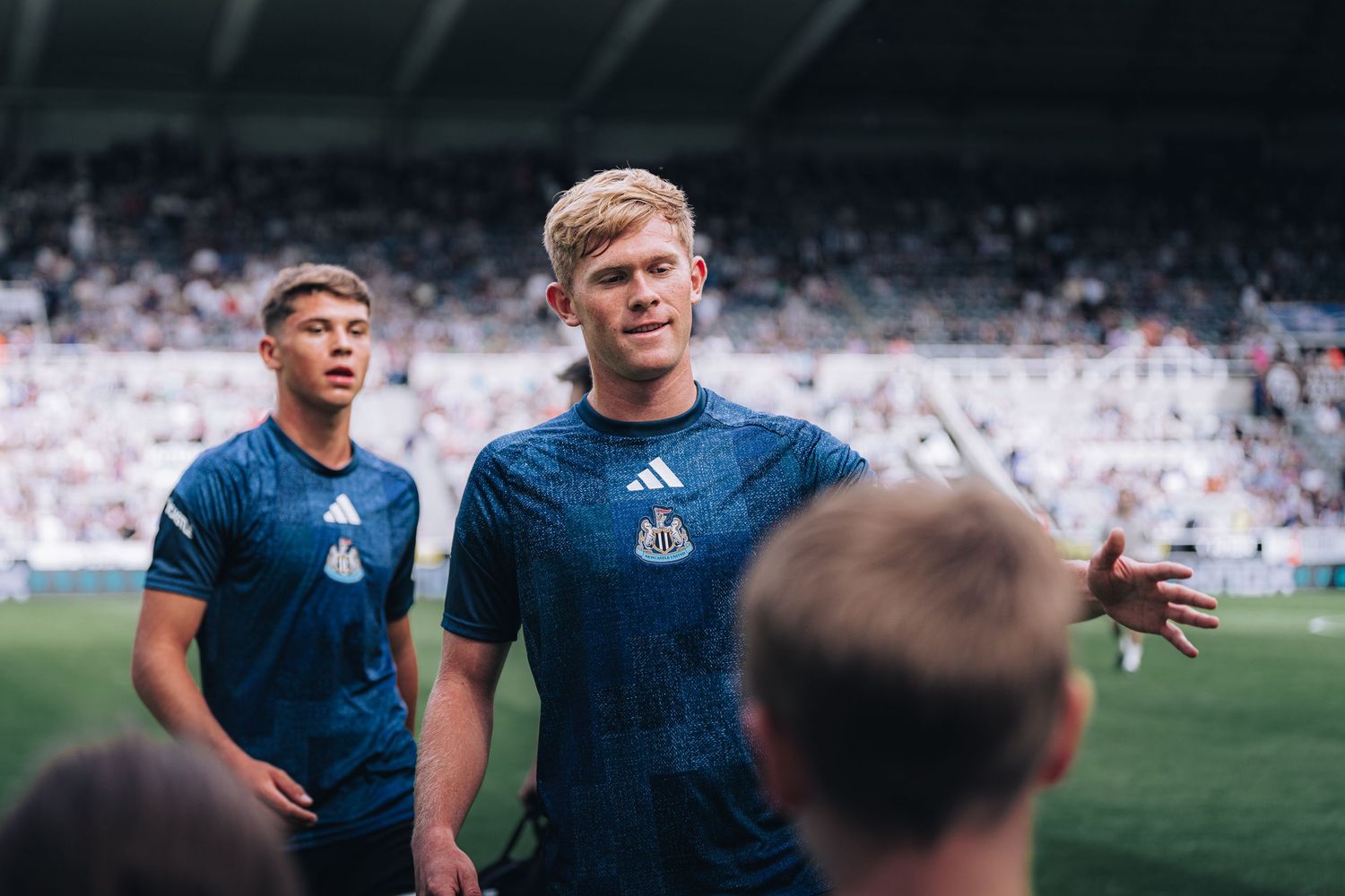 Two Newcastle United players in blue Adidas training kits smiling on the pitch at St. James Park.