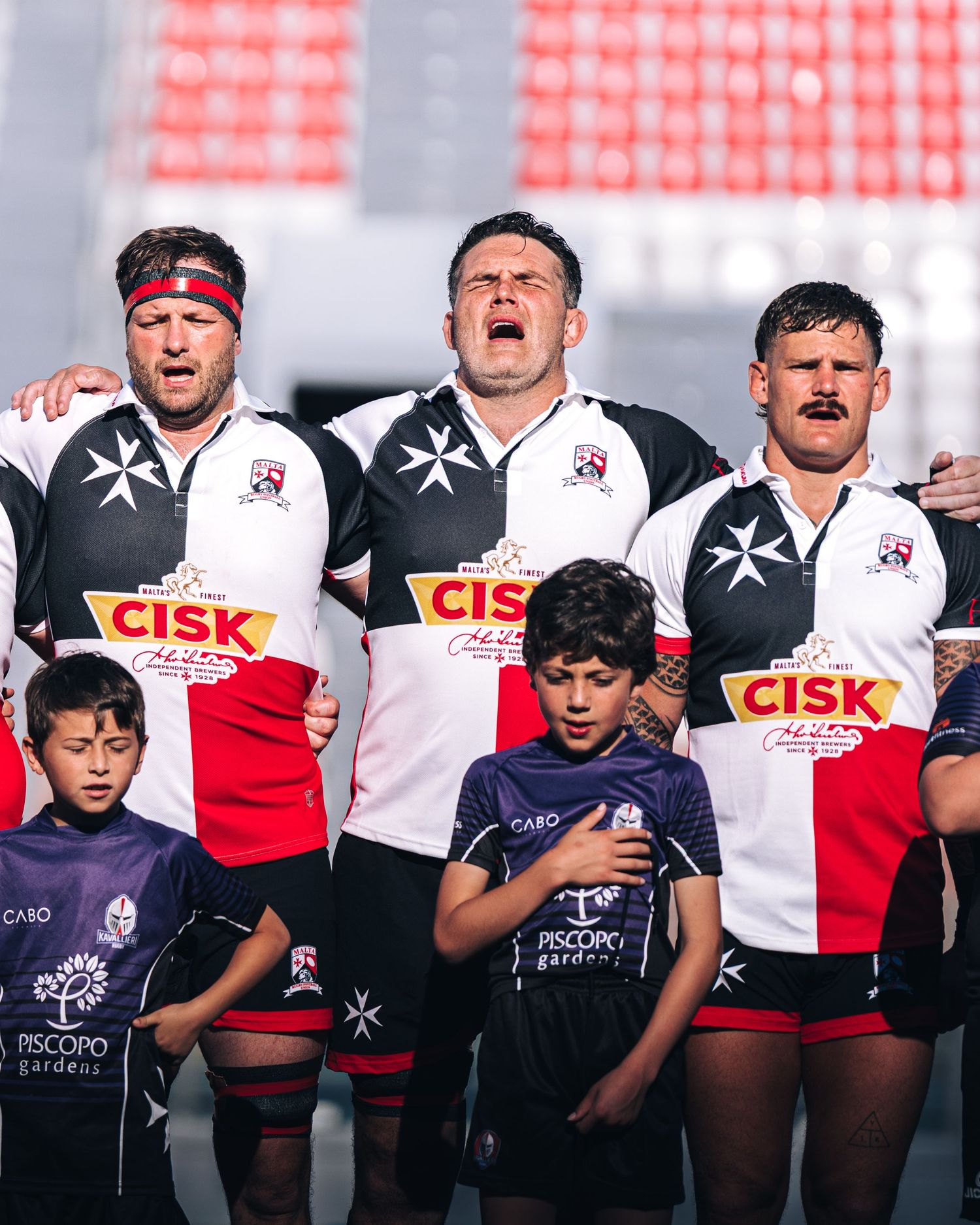 Malta rugby league players singing national anthem with young mascots before international match.
