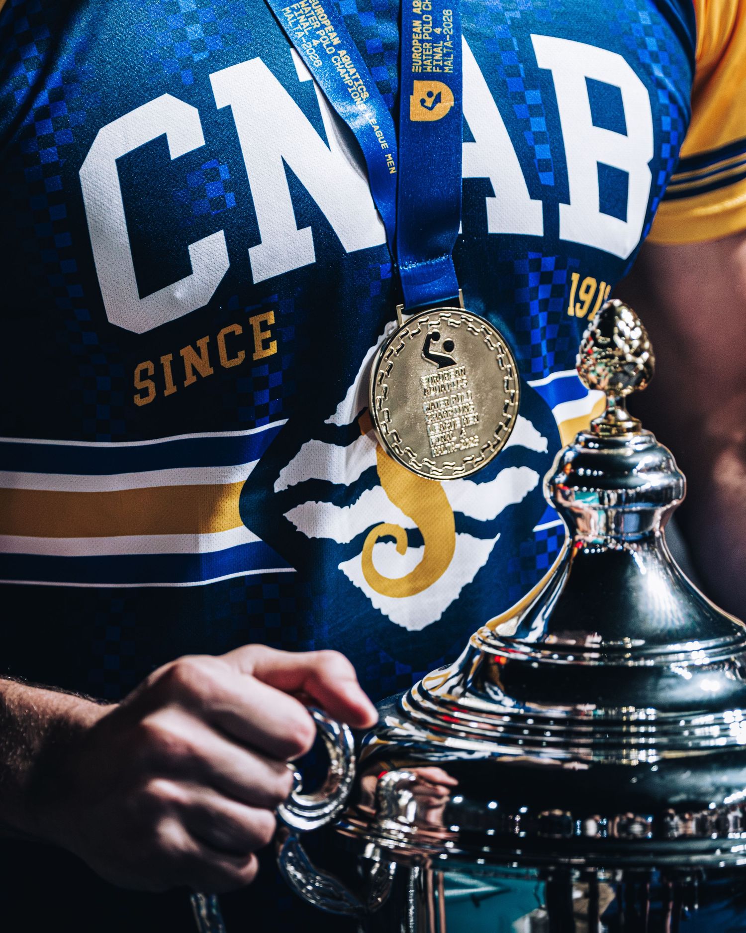 Water polo player displays gold medal and trophy wearing CNAB team jersey.