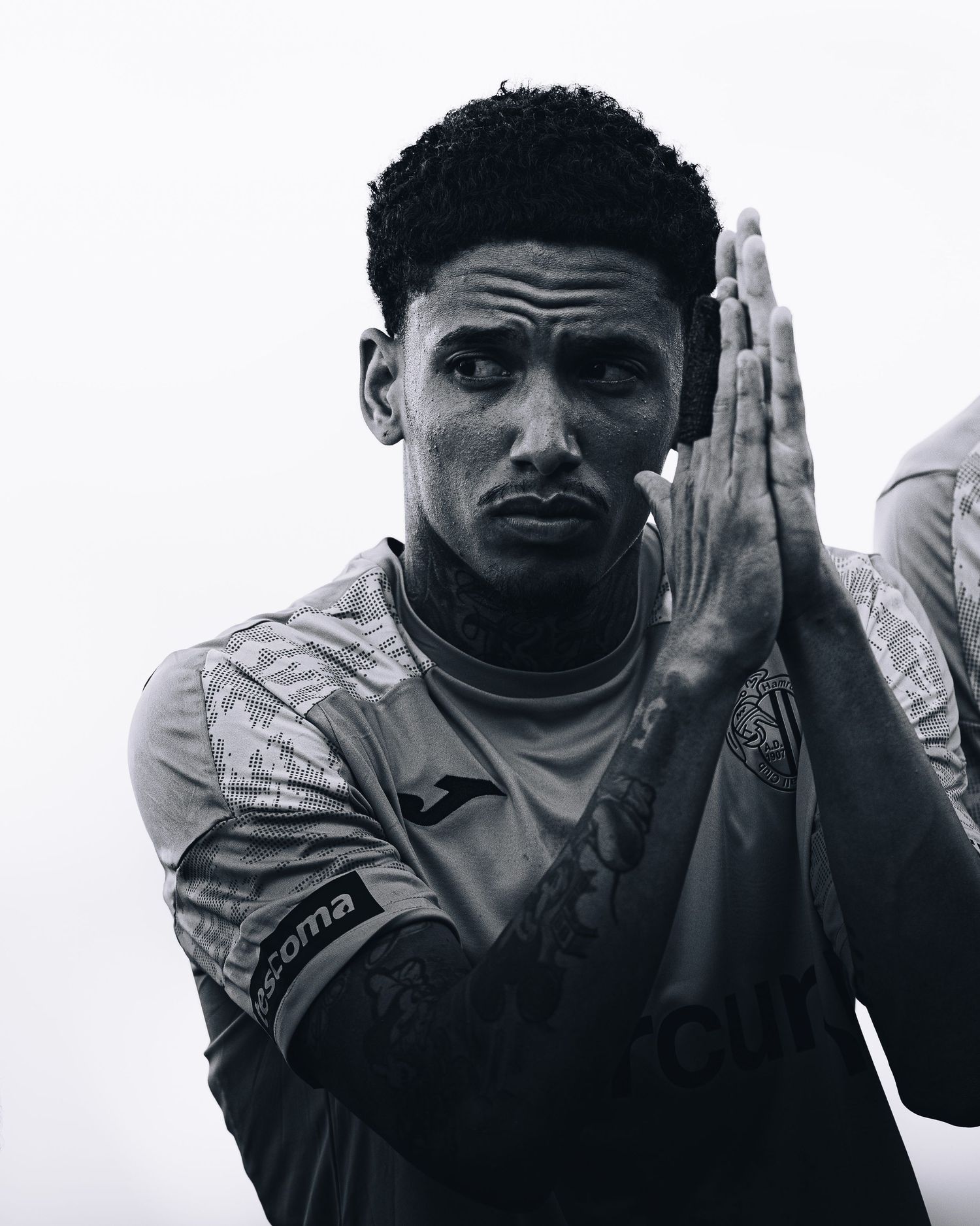 Black and white portrait of tattooed soccer player in Joma kit with hands pressed together.