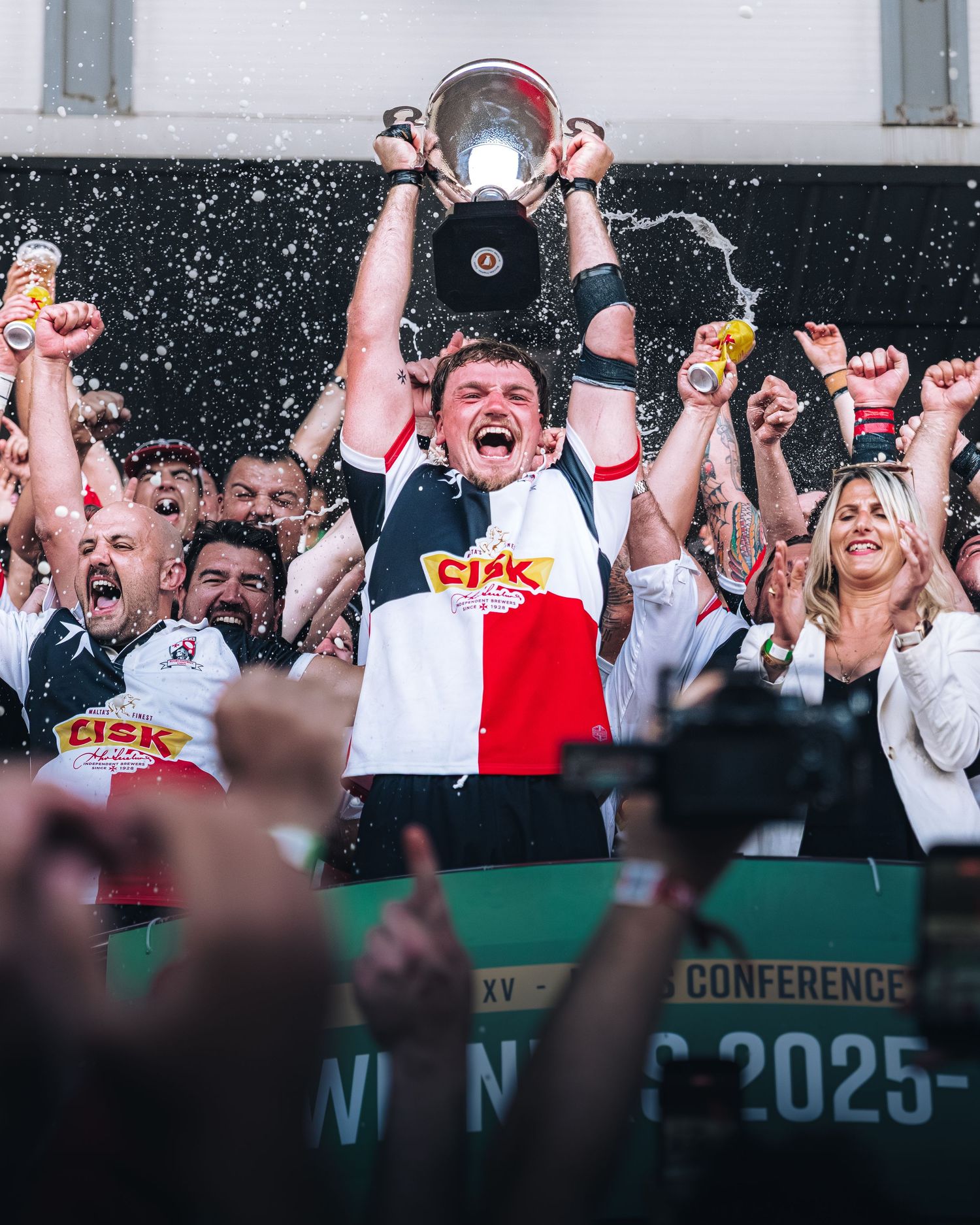 Rugby team captain lifts trophy celebrating conference championship win with cheering teammates.