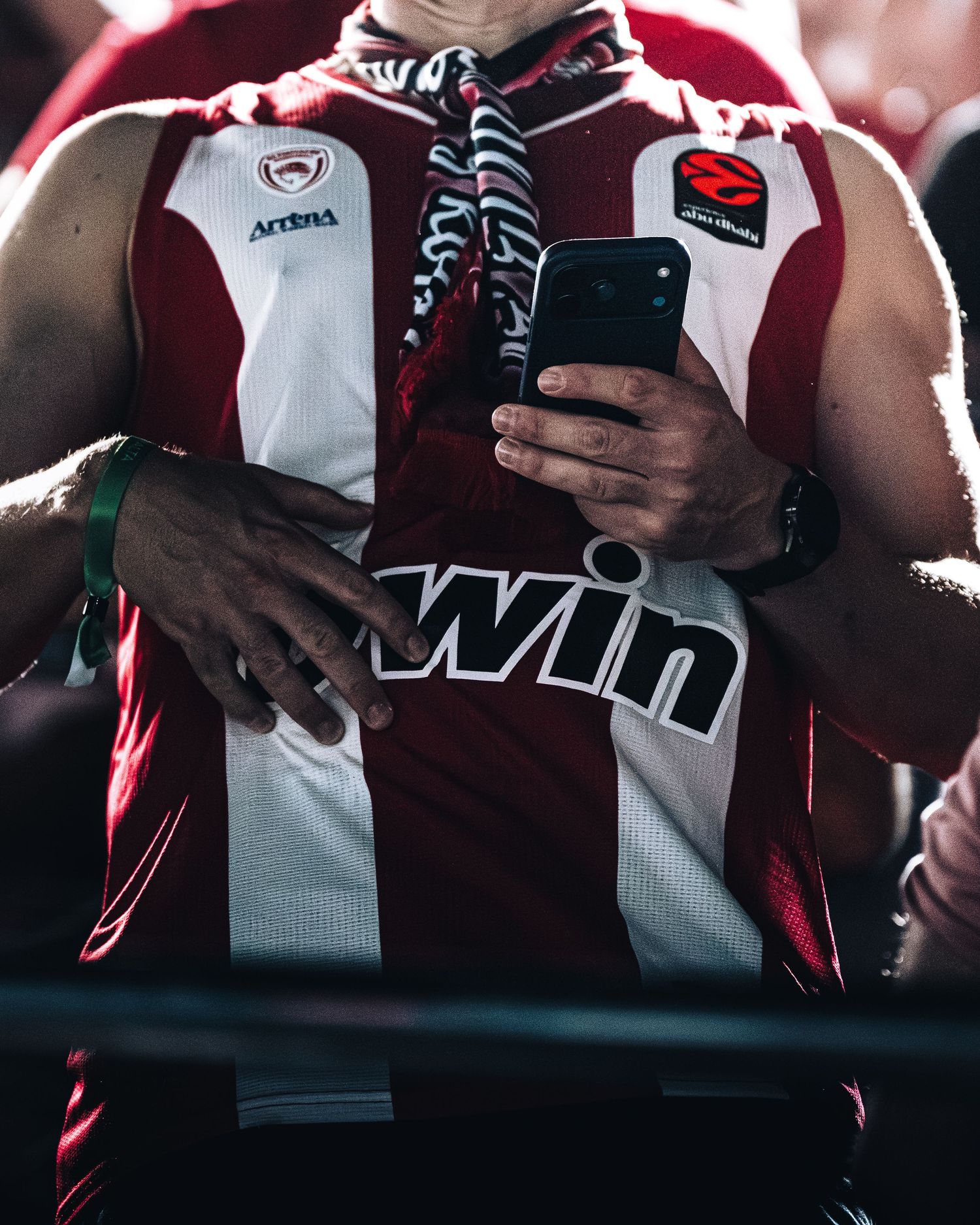 Sports fan in Olympiacos red and white Euroleague basketball jersey uses smartphone at arena.