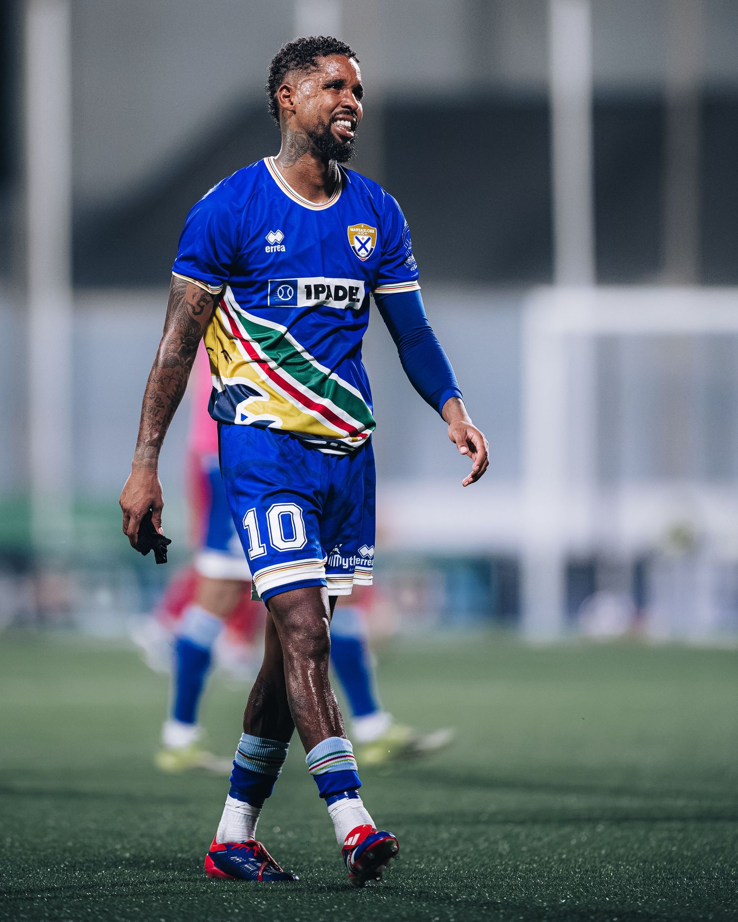Tattooed soccer player in blue number 10 Errea jersey walking on football pitch.
