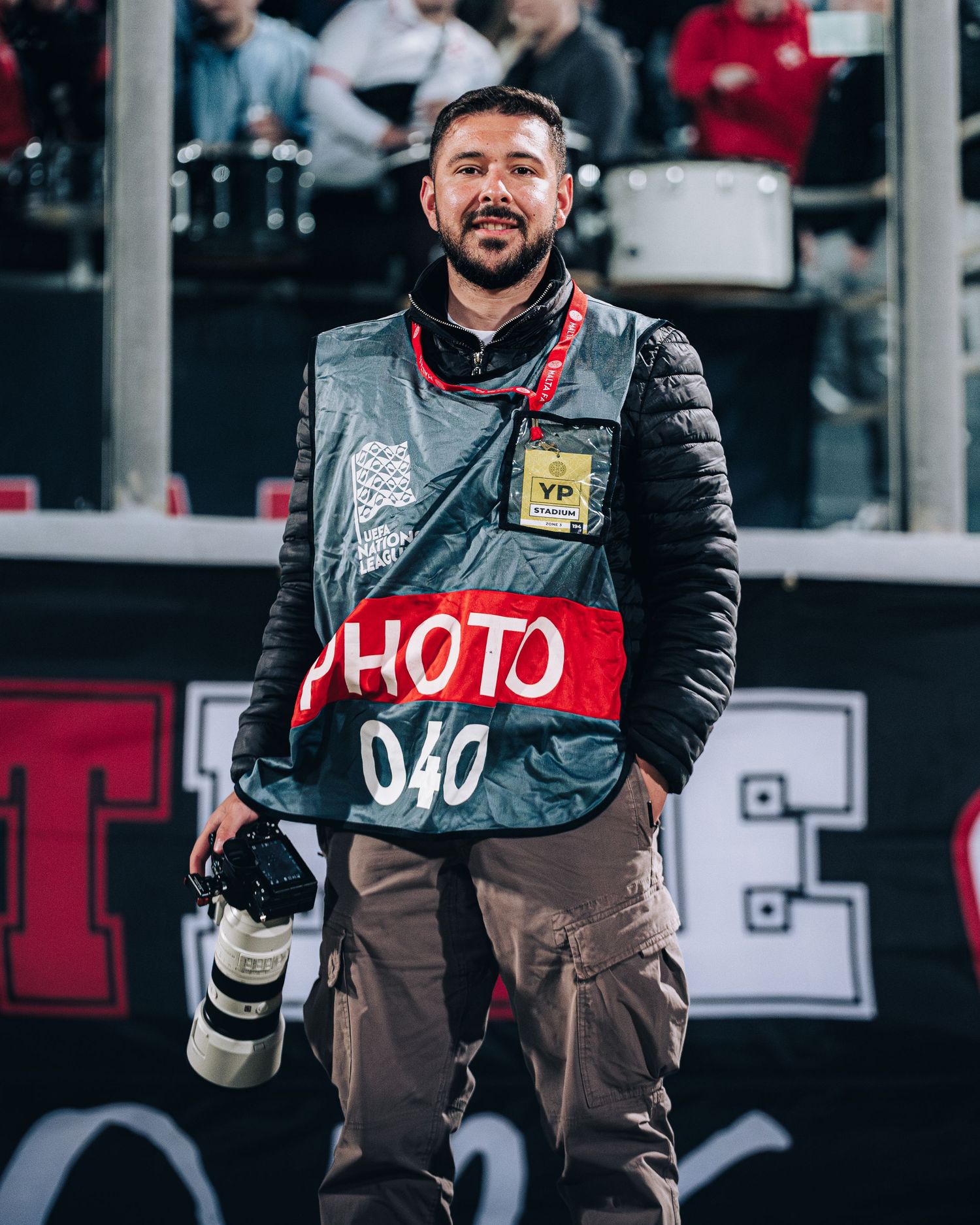 Sports photographer wearing photo vest and holding camera lens on sidelines of stadium.