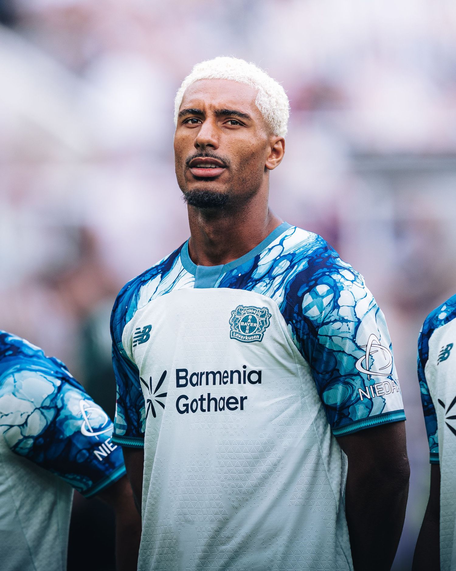 Soccer player in Bayer Leverkusen white and blue Bormenia kit with bleached blonde hair standing on field.
