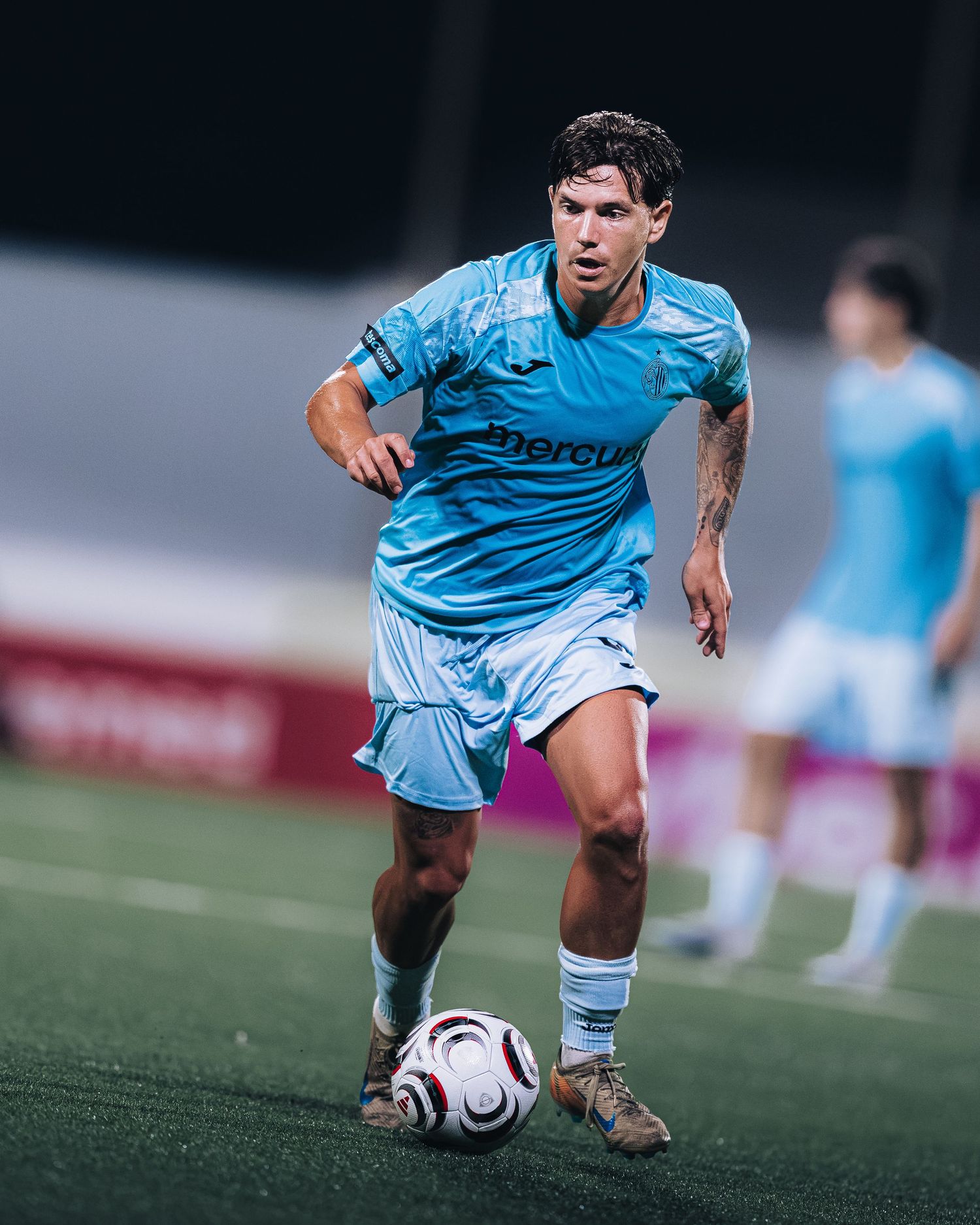 Soccer player in light blue kit dribbling a ball during a night match on a floodlit pitch.