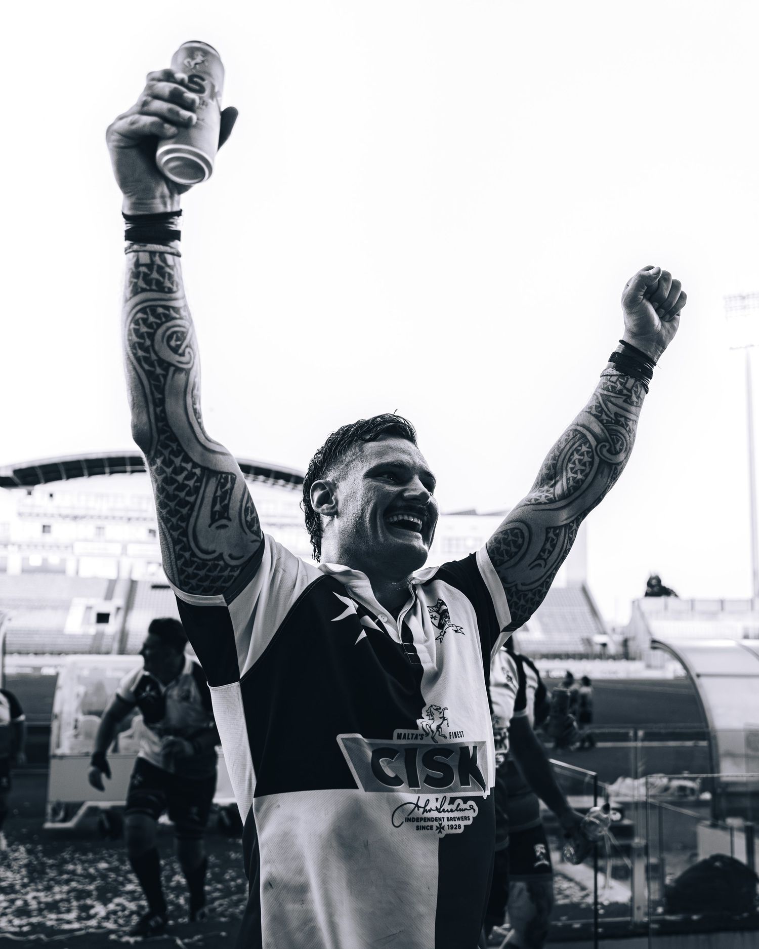 Black and white photo of a tattooed male athlete celebrating with arms raised holding a drink can.