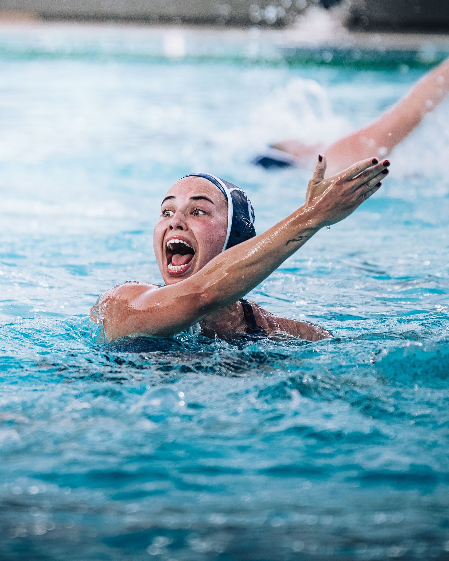 Excited female water polo player in swim cap celebrates with open mouth and raised arm in pool.