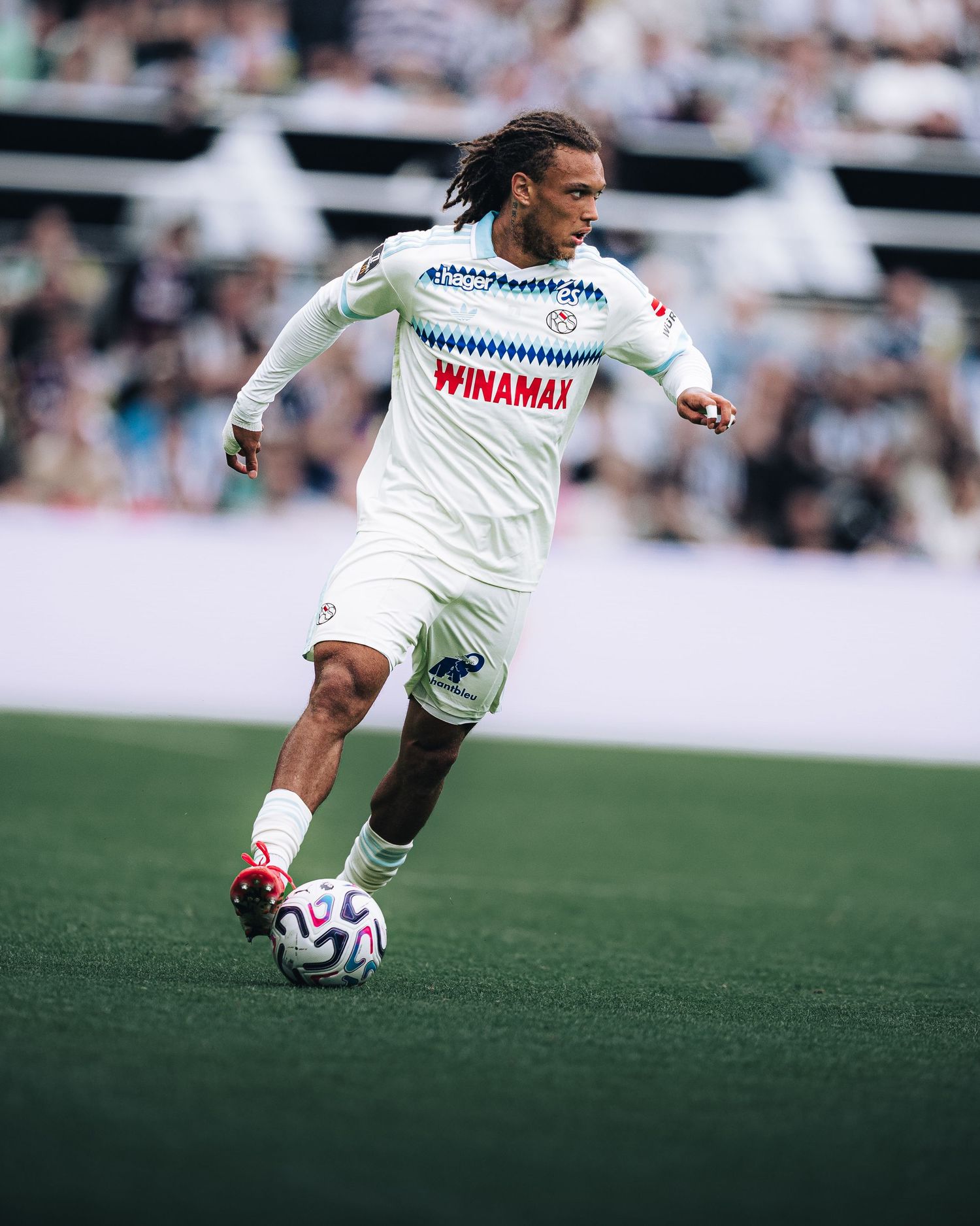 Footballer in white Winamax kit dribbles ball on green pitch during match.