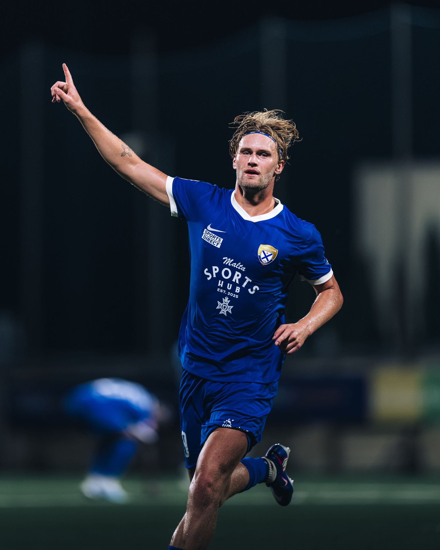 Soccer player in blue Nike jersey celebrates goal with finger pointing skyward at night.