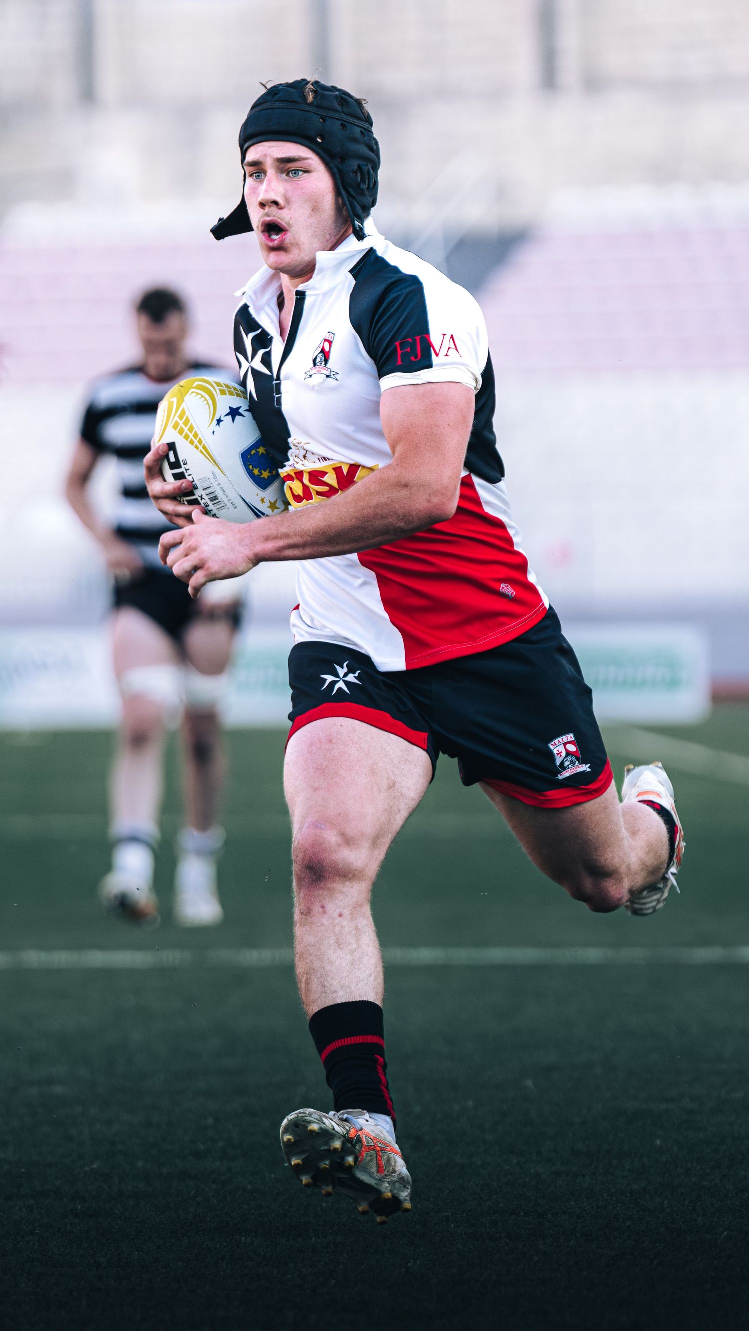Young rugby player wearing headguard sprinting with ball during competitive match in stadium.