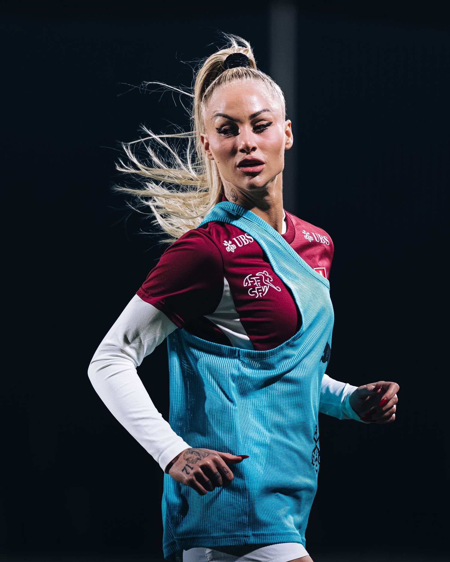 Female footballer with blonde ponytail running in maroon and teal training kit during practice.